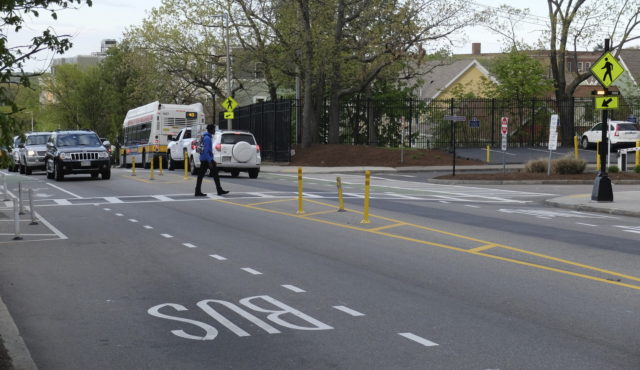 A young man crosses the street at a mid-block crossing, where the street has been narrowed using temporary bollards and other quick-build infrastructure to narrow the road and reduce vehicle speeds. A bus lane is also present along the curb for each side of the street.