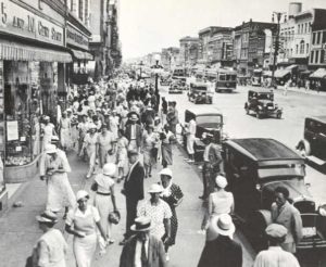 A photo of Broad Street in Richmond VA circa 1925, where many people are walking on a wide siewalk, with cars and streetcars in a broad boulevard along a humming retail cooridoor.