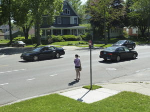 A woman waits to cross the street at an uncontrolled, unmarked crosswalk at an intersection in Saint Paul. No one is yielding to her.