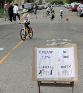 Photo of sign saying "Oakland Slow Street No Thru Traffic" while families stand and play in the street behind it