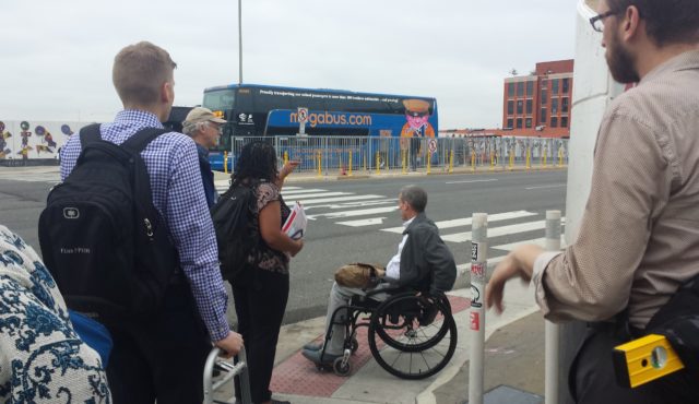 A group of people look out at a crosswalk. One of the people is a in a wheelchair and another is using a walker.