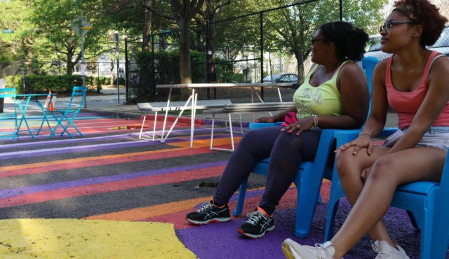 Header Image: Two Black women sit in lawn chairs in parking lot painted with colorful designs and set up with outdoor seating