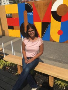 ROsie Jaswal sitting on a bench on Jaspar Ave, Edmonton; colorful mural behind her.