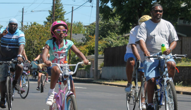 A Black girl rides her bike down the street, accompanied by two older Black men, also on bicycles.