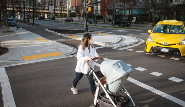 Woman crosses the street with a baby carriage in Vancouver, BC. A taxi is stopped as it turns onto the street, waiting for her to cross. The woman is looking at the taxi.