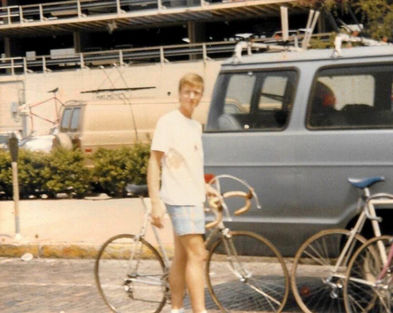Young man standing next to his bike with a blue car in the background.