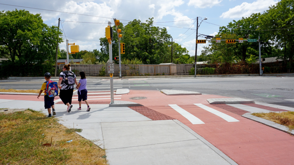 A mother with children uses a crosswalk with a dedicated bike lane and pedestrian signals, implemented as part of infrastructure improvements for the Austin Safe Routes to School program.