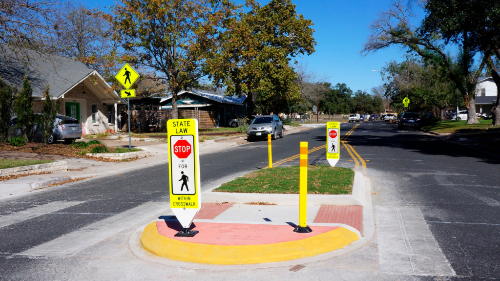 A pedestrian refuge island with signs indicating "State Law: Stop for Pedestrians," implemented as part of an infrastructure improvement for the Austin Safe Routes to School program in a residential neighborhood.