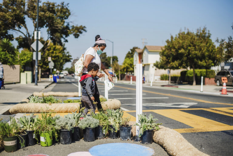 A mother and son walk across a temporary curb extension comprised of straw wattles and potted plants during the tactical urbanism demonstration project for the San Pablo SR2S Plan.