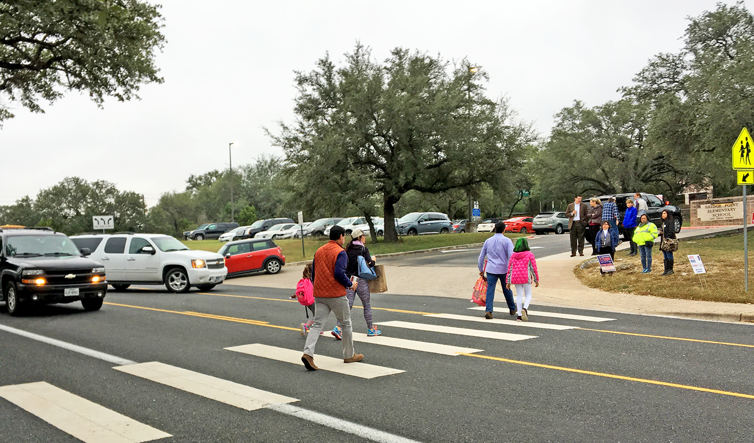 Children and adults use a crosswalk on a busy street near a school, with cars waiting as they cross.