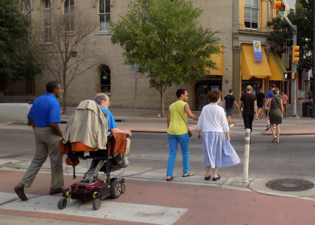 A diverse group of pedestrians, including a person in a wheelchair, cross a street with a designated bike lane and bollards.