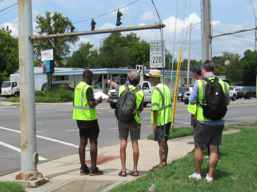 A group of people in safety vests stand at a sidewalk near a busy intersection, conducting a walk audit.