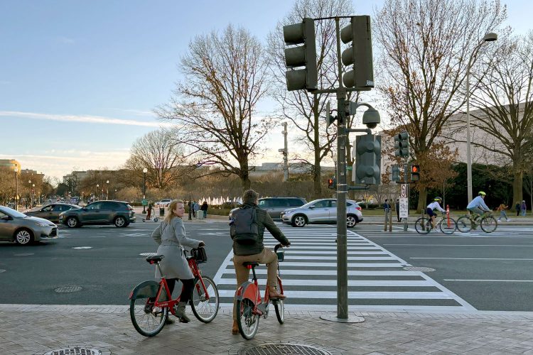 Two people wait at a crosswalk with Capital Bikeshare bikes.